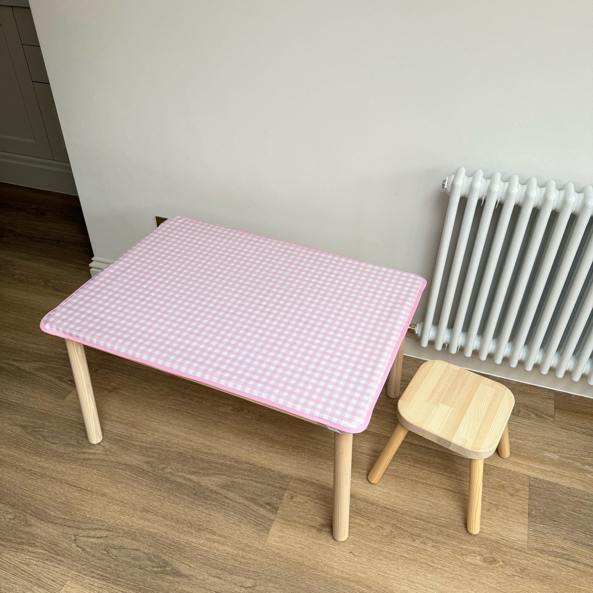 Small wooden table with a pink checkered cover next to a wooden stool on a wooden floor.