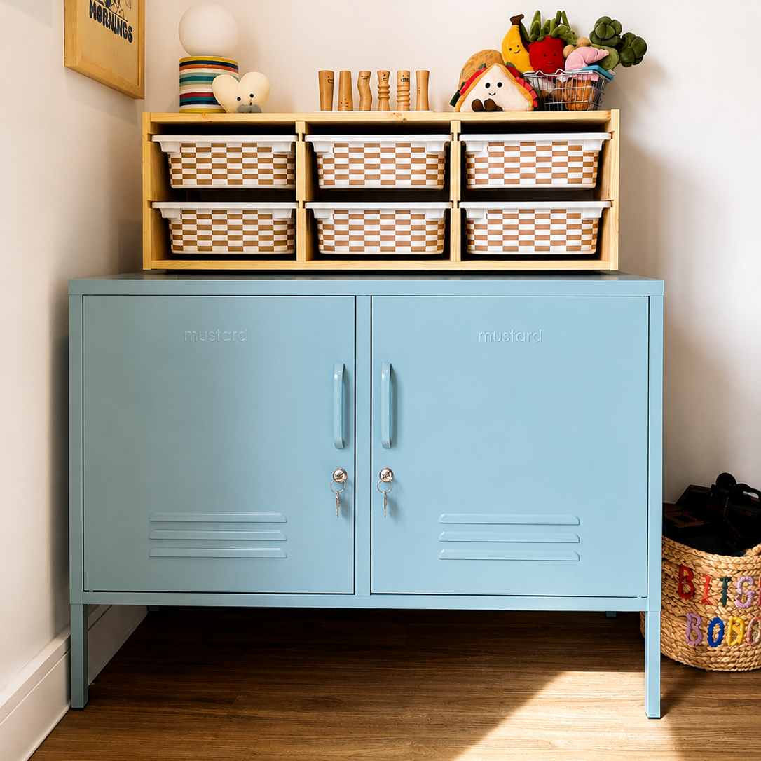 A light blue metal storage cabinet with two doors stands on a wood floor against a white wall. On top of the cabinet is a Trofast shelf holding six woven storage baskets, toys, and small decor items.