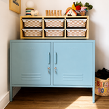 A light blue metal storage cabinet with two doors stands on a wood floor against a white wall. On top of the cabinet is a Trofast shelf holding six woven storage baskets, toys, and small decor items.