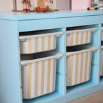 Children's play area with a blue storage unit and striped bins, featuring toys and a dollhouse.