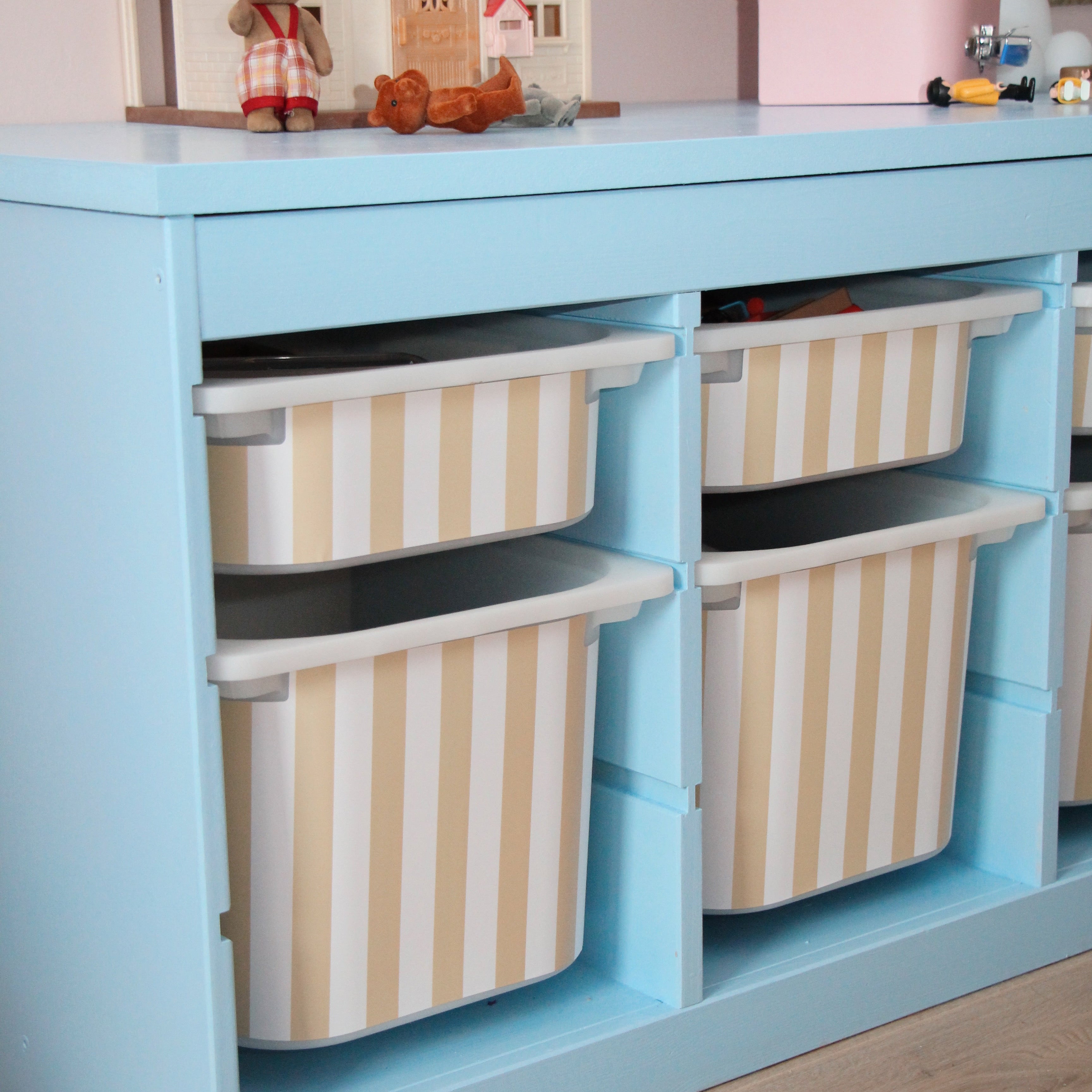 Children's play area with a blue storage unit and striped bins, featuring toys and a dollhouse.