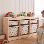 A light natural wood children's storage cabinet with six alphabet-print fabric bins, styled in a bright nursery with toys and a child sitting nearby.