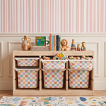 A light wood children's storage organizer with six pastel checkered bins filled with toys, styled in a nursery setting with pink striped wallpaper, a bookshelf, and a rug.