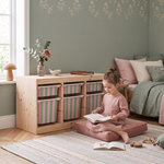A young girl sits on a pink cushion on the floor, reading a book in a cozy, styled children's bedroom. To her left is a light wood storage cabinet with six pink and green striped fabric bins.